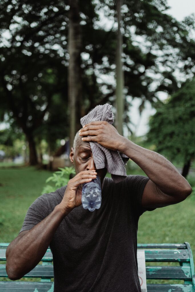 A man wipes sweat with a towel while hydrating in a lush outdoor setting.